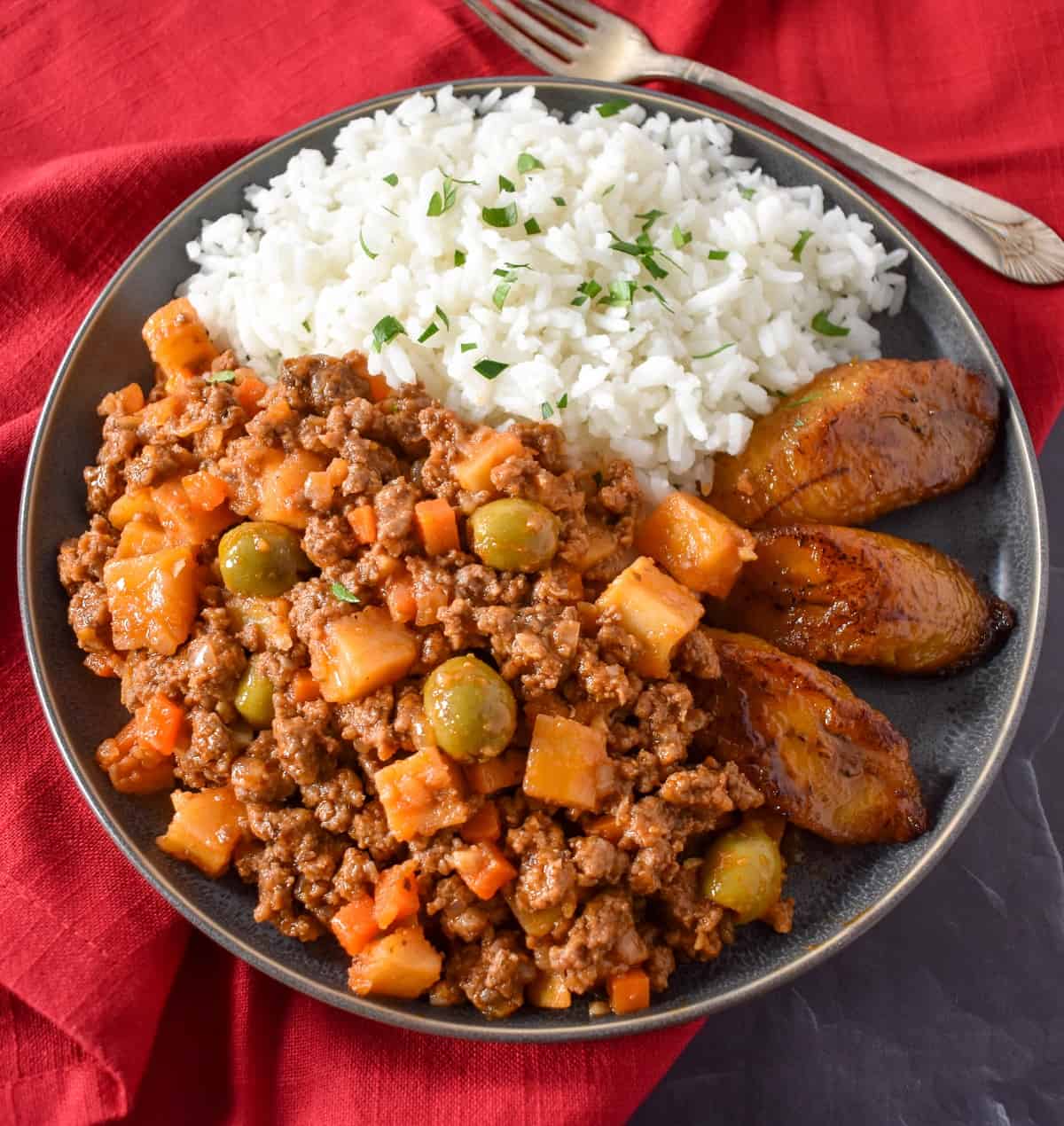 Close up image of the finished picadillo served with white rice and fried sweet plantains on a gray plate.