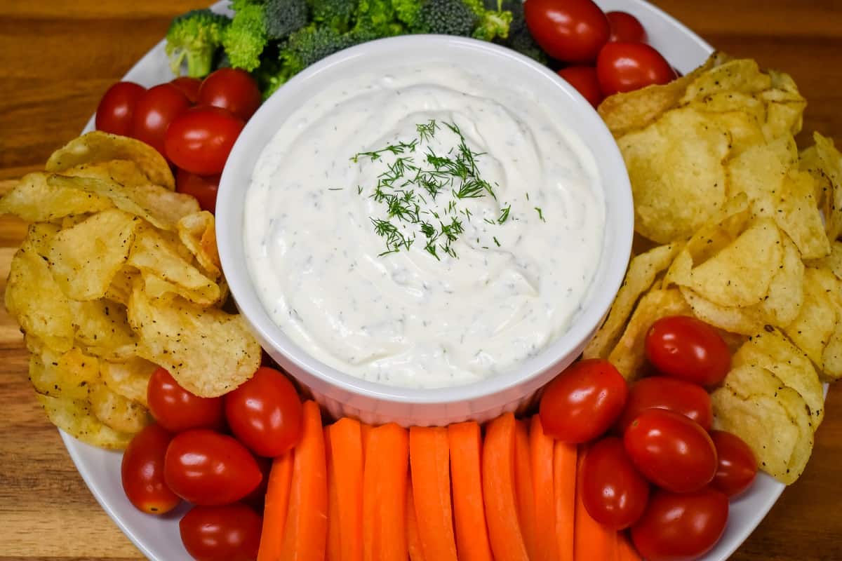 A top picture of dill dip served in a large white bowl with chips, grape tomatoes, carrots and broccoli florets arranged around the bowl.