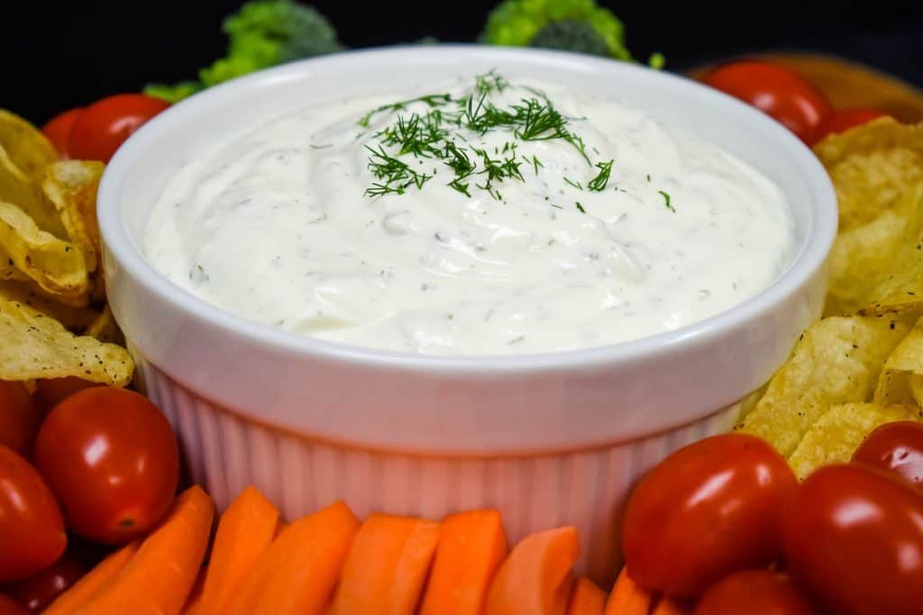 A close up picture of dill dip served in a large white bowl with chips, grape tomatoes, carrots and broccoli florets arranged around the bowl.