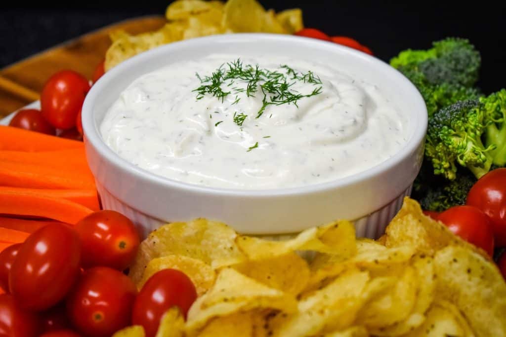 Dill dip served in a large white bowl with chips, grape tomatoes, carrots and broccoli florets arranged around the bowl.