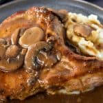 A thick pork chop covered with brown mushroom gravy served on mashed potatoes on a gray plate.