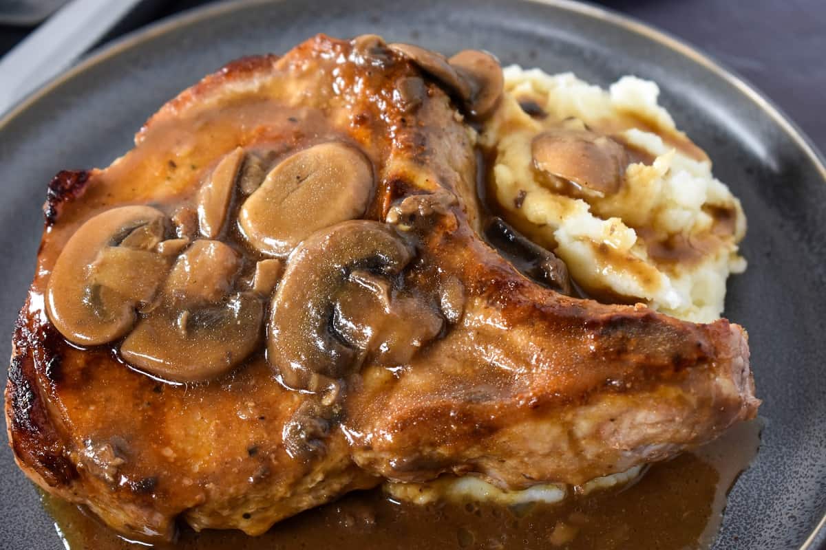 A thick pork chop covered with brown mushroom gravy served on mashed potatoes on a gray plate.