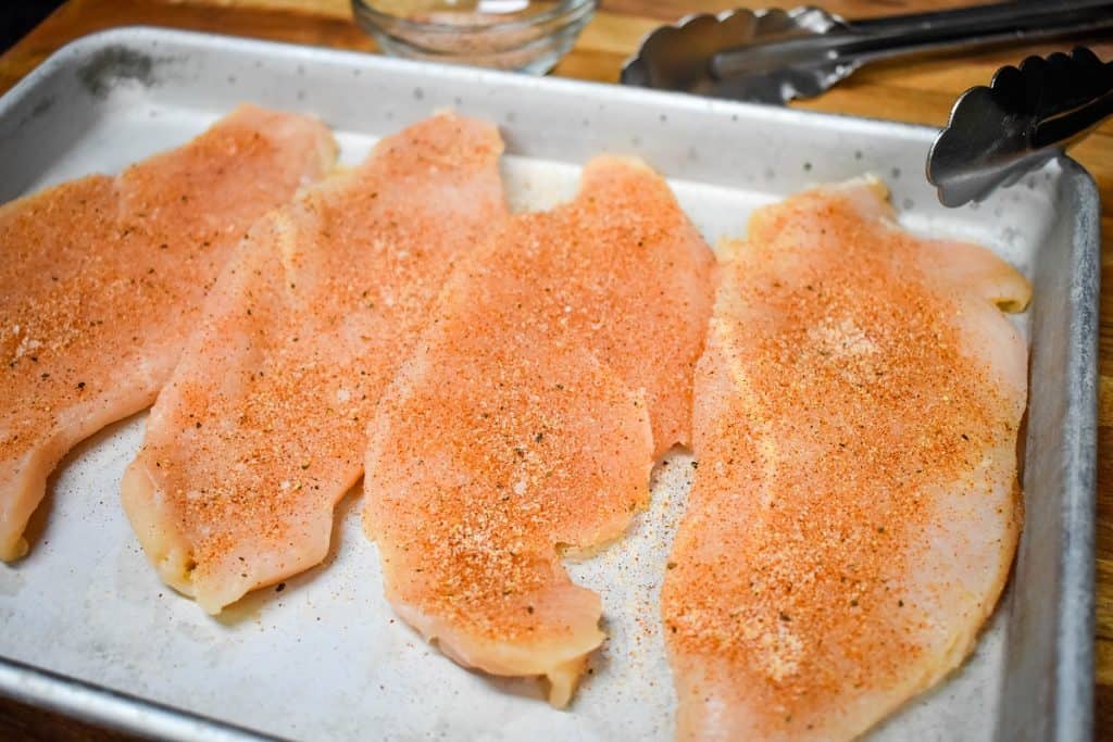 Seasoned, thin chicken breasts on a metal baking sheet.