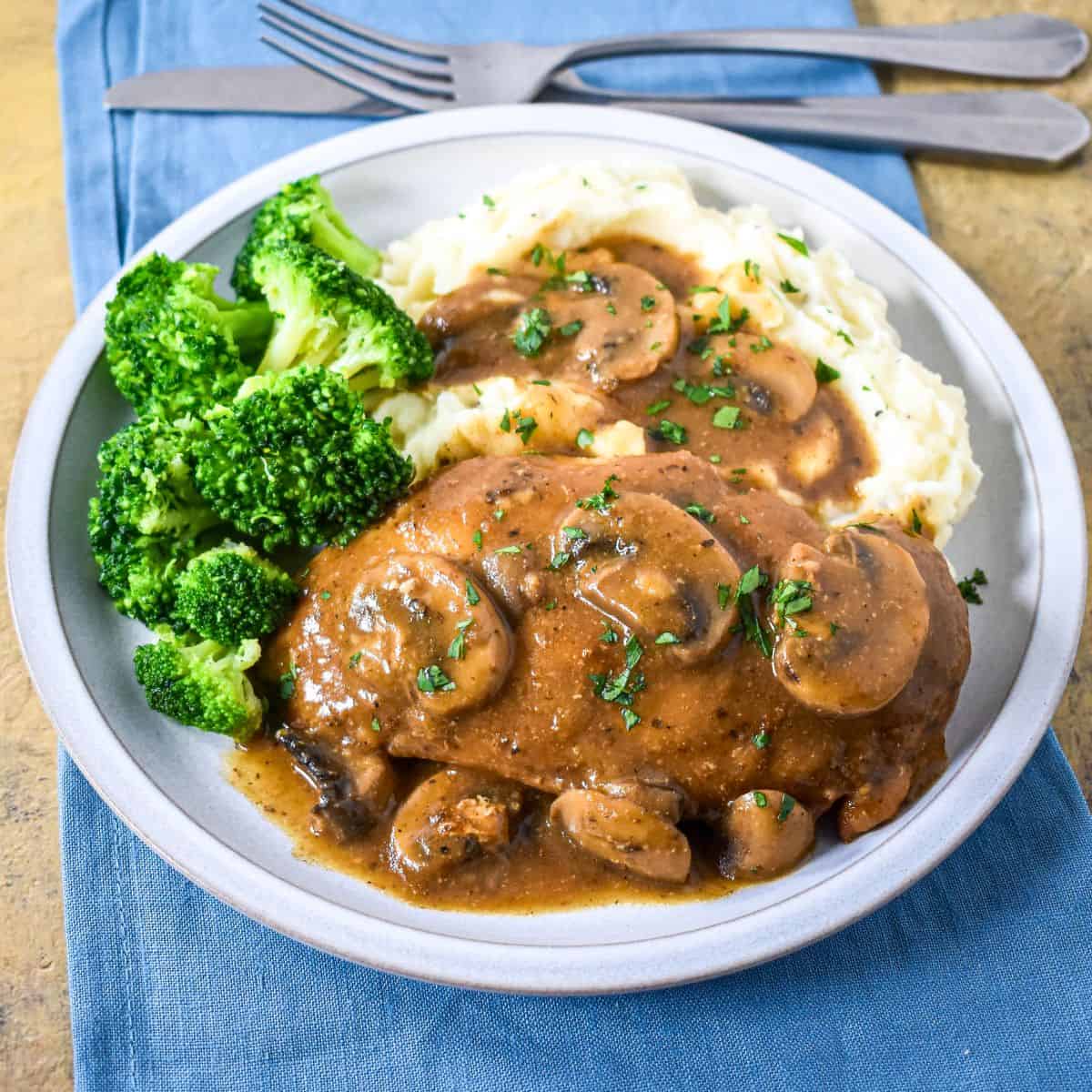 Chicken and mushrooms with brown gravy served with mashed potatoes and broccoli served on a white plate.