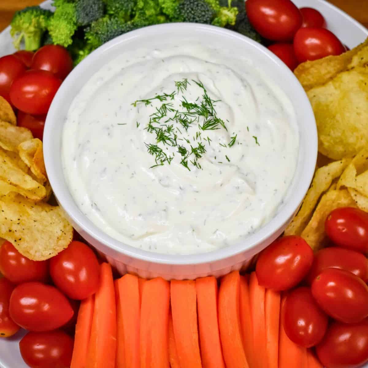 Fresh dill dip served in a white bowl surrounded with vegetables and chips