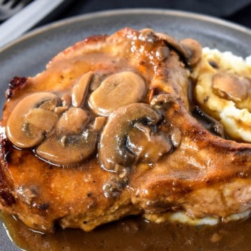 A thick pork chop covered with brown mushroom gravy served on mashed potatoes on a gray plate.