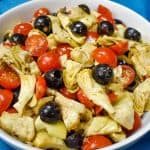 A cold salad with quartered artichokes, grape tomatoes, black olives and sliced onions tossed with an oil and vinegar dressing and served in a large white bowl on a blue cloth.