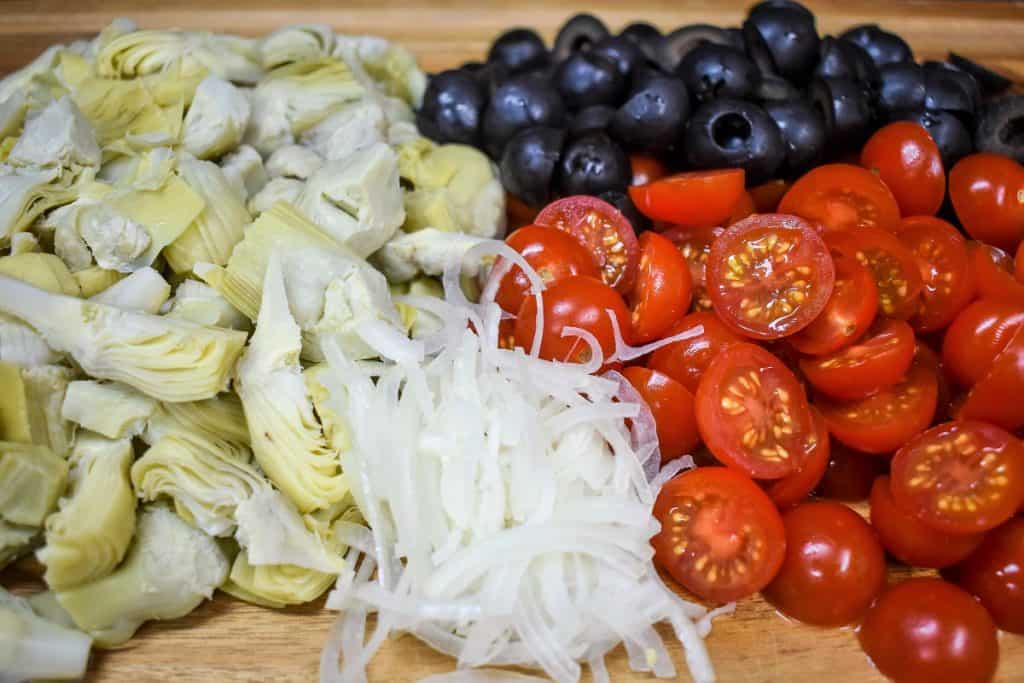 Quartered artichokes, grape tomatoes cut in half, black olives and thinly sliced onions displayed on a wood cutting board.