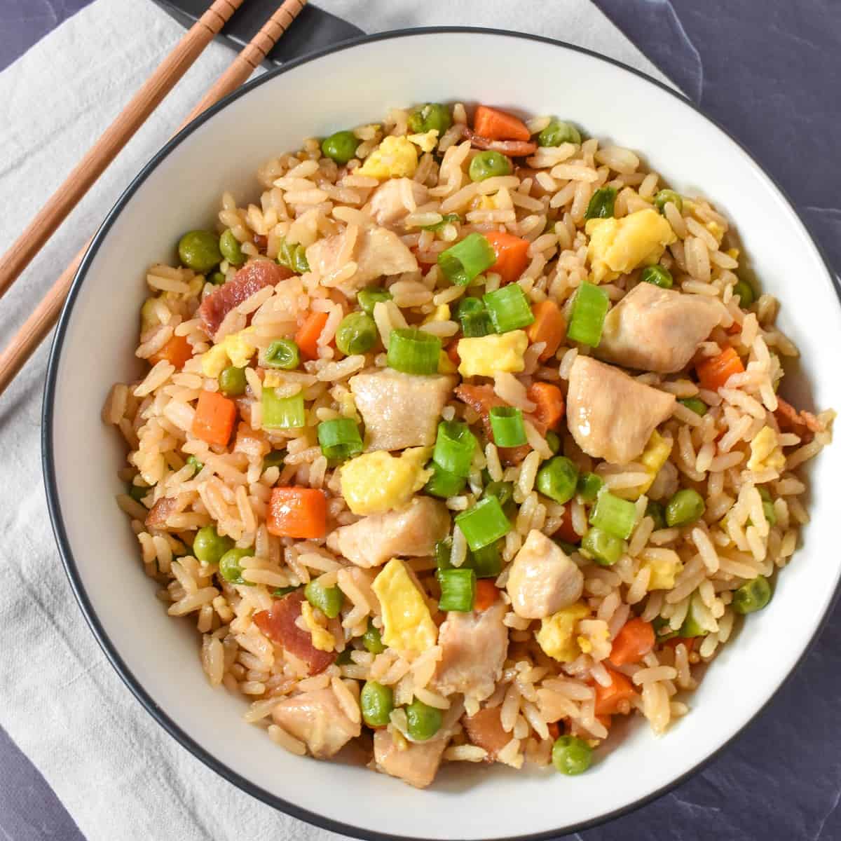 An image of the chicken fried rice served in a white bowl with a black rim. The bowl is set on a beige linen on a black table and there are chopsticks to the top, left side.