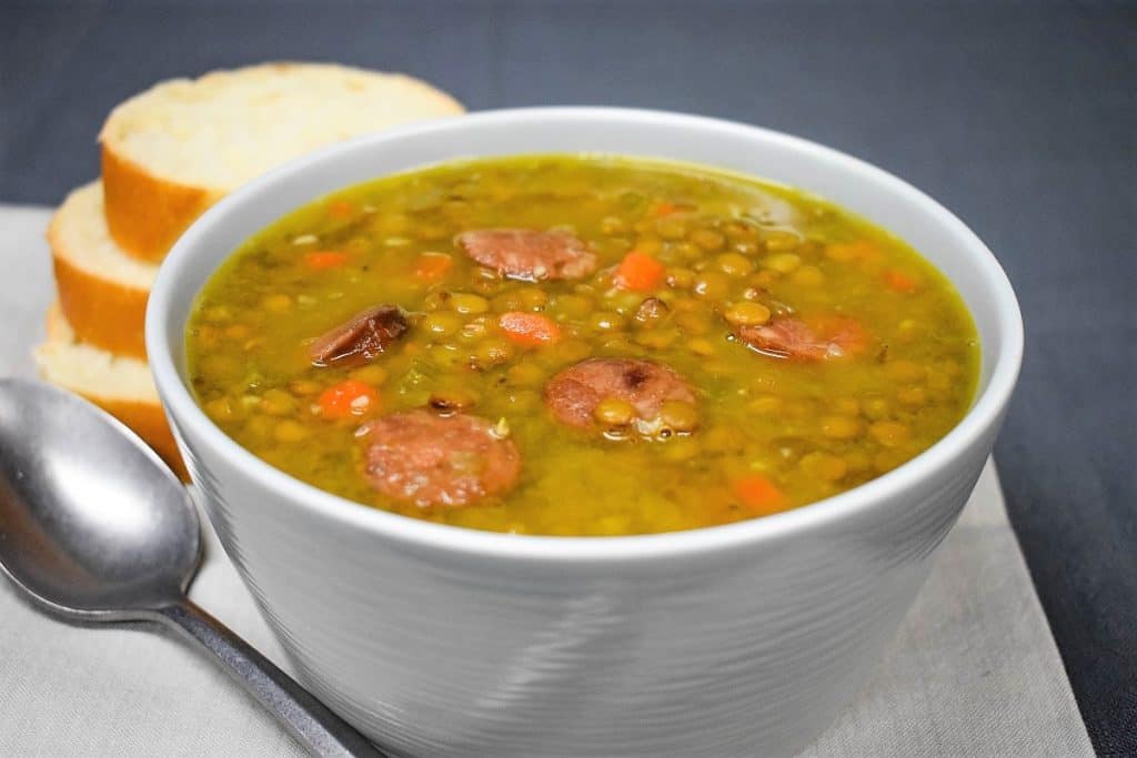 Lentils with andouille sausage soup served in a light gray bowl with bread slices in the background.