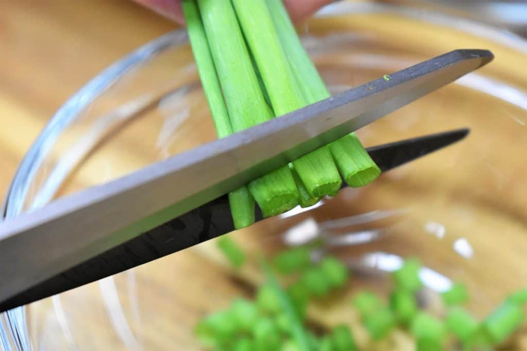 Green onions being snipped by kitchen shears into a clear bowl.