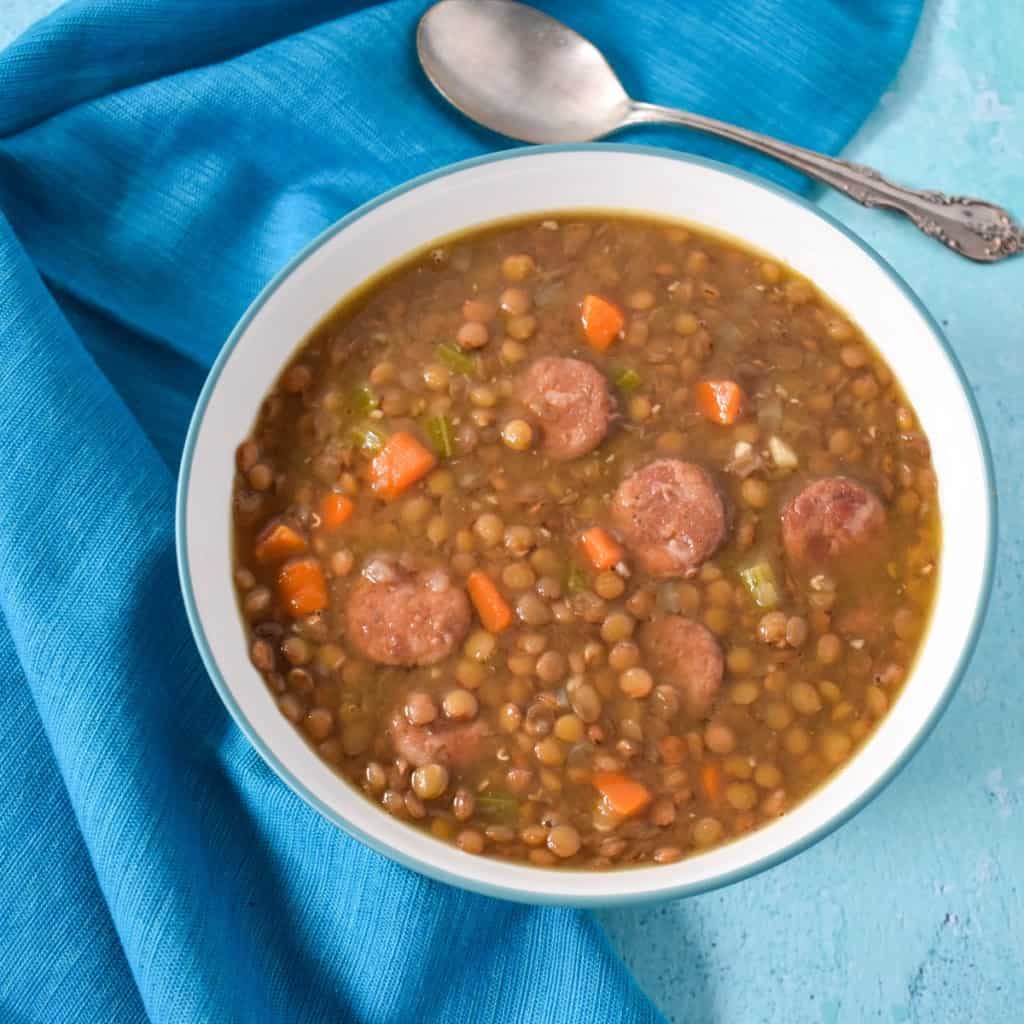 The lentil and andouille sausage soup served in a white bowl with an aqua rim with an aqua linen and a spoon to the top, right side.