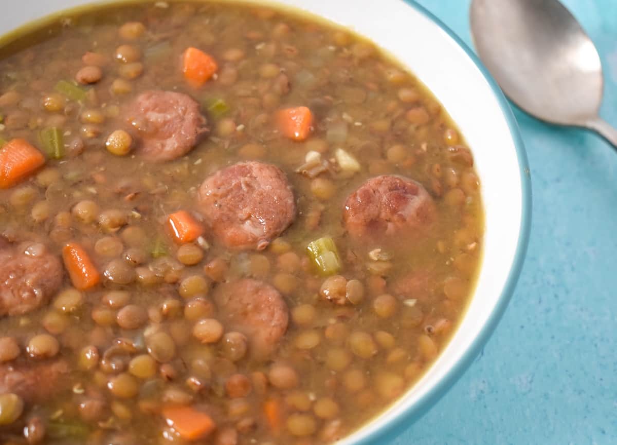 A close up image of the lentil andouille soup served in a white bowl with an aqua rim, set on a light blue table.