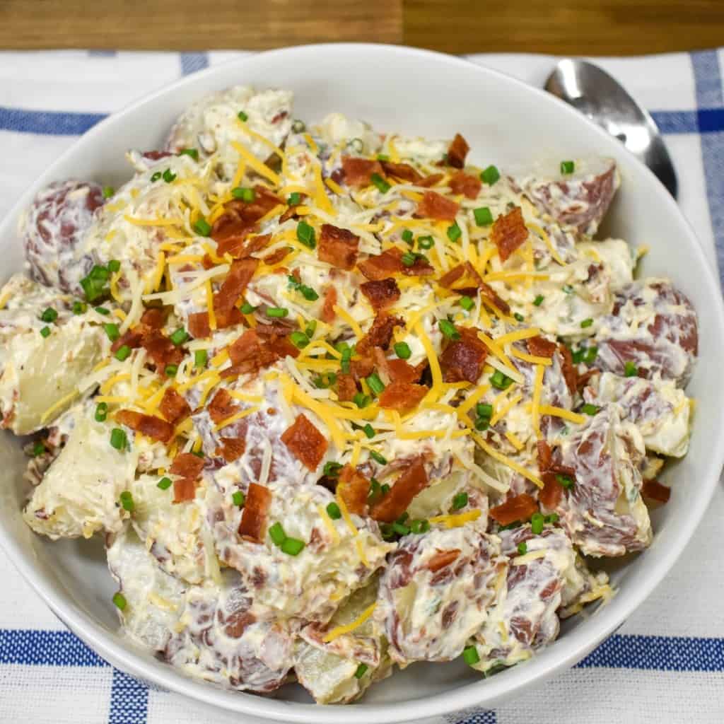 An image of loaded potato salad served in a large white bowl and garnished with bacon, cheese, and chives.
