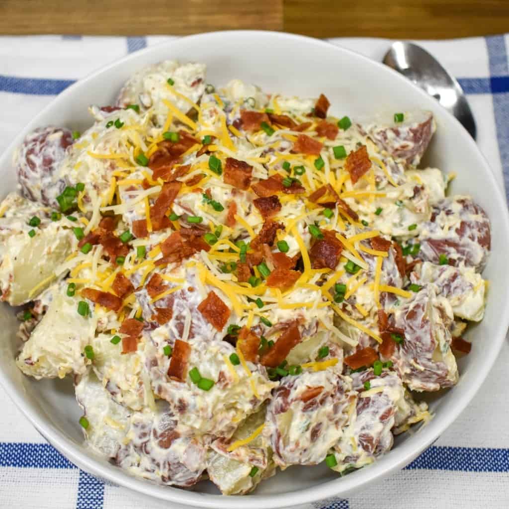 An image of loaded potato salad served in a large white bowl and garnished with bacon, cheese, and chives.