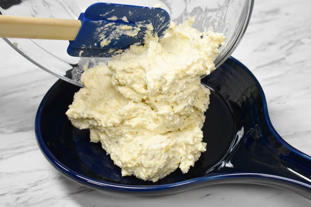 Artichoke dip mixture being transferred from a glass bowl to a blue baking dish.