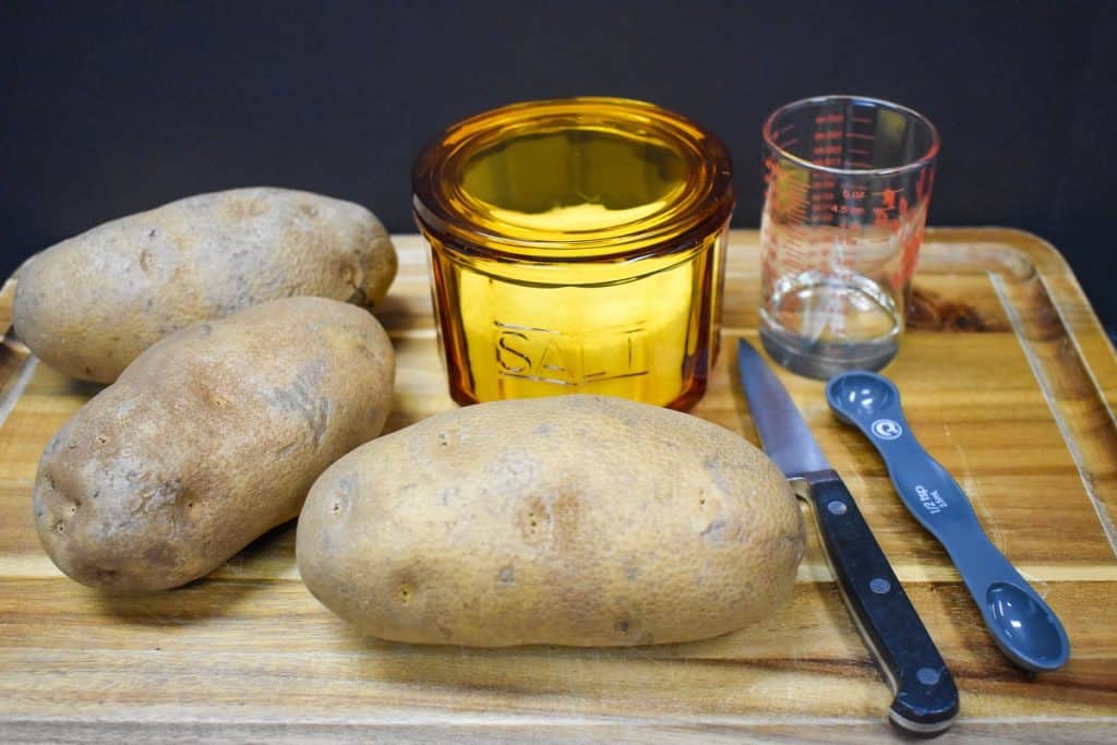 Three large russet potatoes, a salt cellar, oil and a paring knife displayed on a wood cutting board.