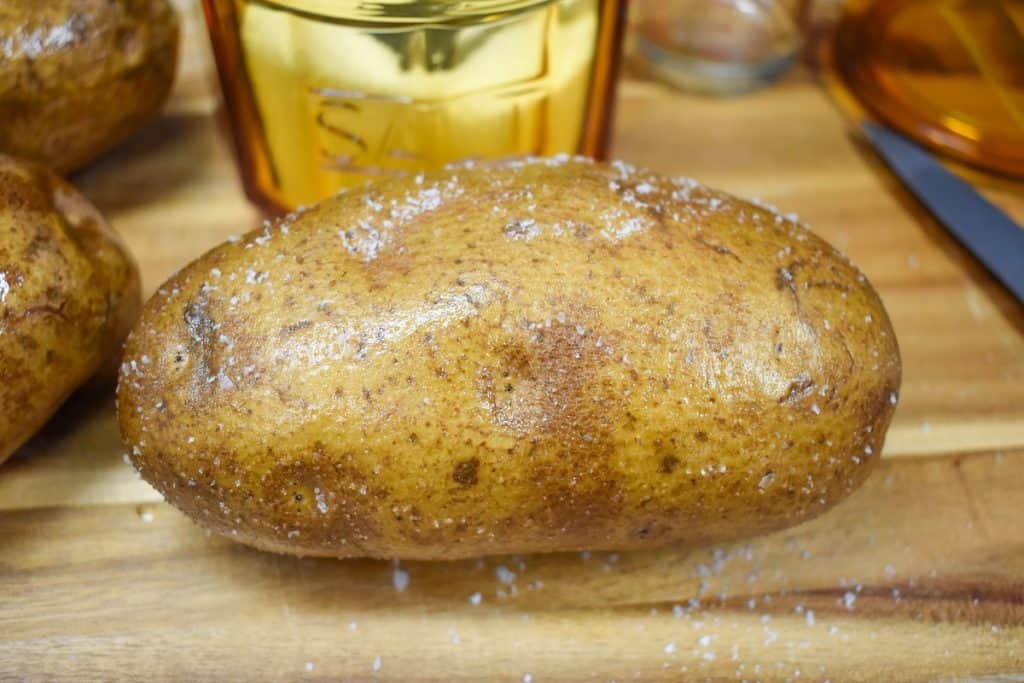 A large russet potato rubbed with oil and salt with an amber colored salt cellar in the background.