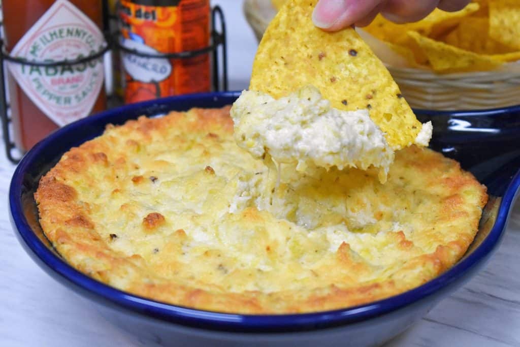 A tortilla chip scooping up some hot artichoke dip with hot sauce in the background.