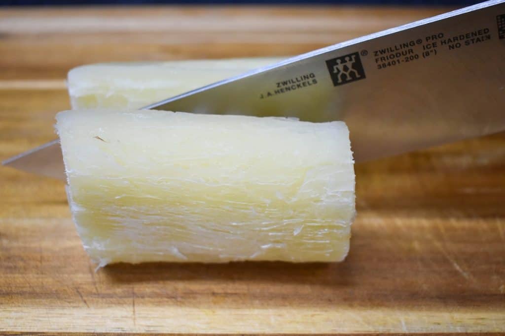 A piece of cooked yuca being cut in half by a large knife on a wood cutting board.