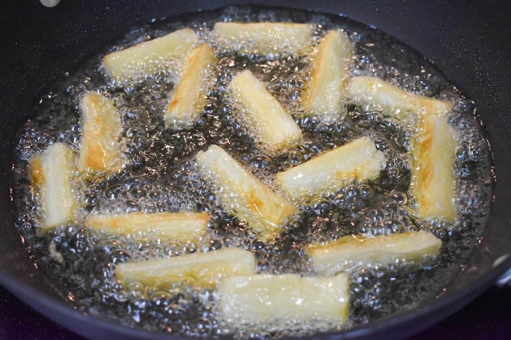 Yuca pieces frying in a large, black skillet. They are turning a soft golden color.