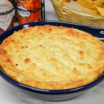 Hot artichoke dip in a blue baking dish with hot sauce and chips in the background.