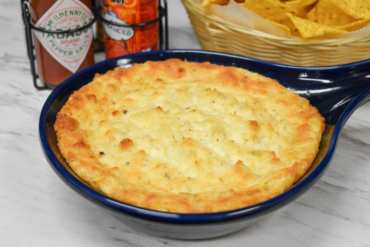 Hot artichoke dip in a blue baking dish with hot sauce and chips in the background.