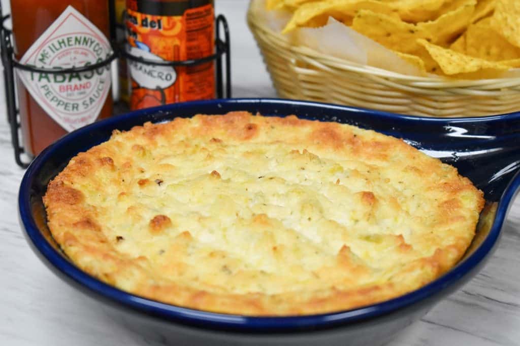 Hot artichoke dip in a blue baking dish with hot sauce and chips in the background.