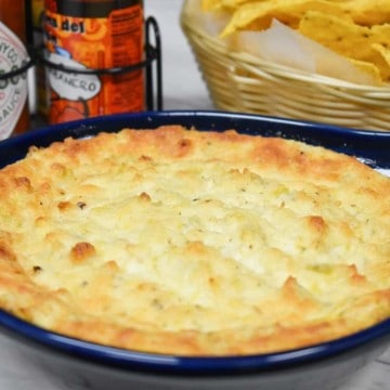 Hot artichoke dip in a blue baking dish with hot sauce and chips in the background.