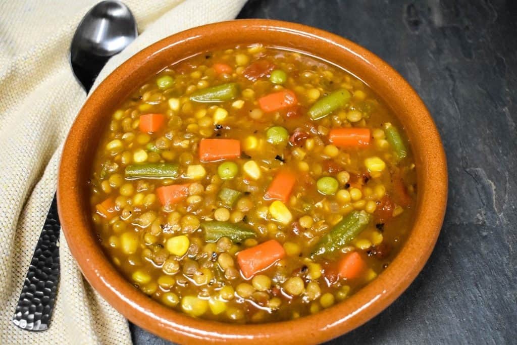 Lentil vegetable soup served in a red clay bowl. Displayed on a slate surface with a beige napkin and spoon on the side.