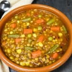 Lentil vegetable soup served in a red clay bowl. Displayed on a slate surface with a beige napkin and spoon on the side.