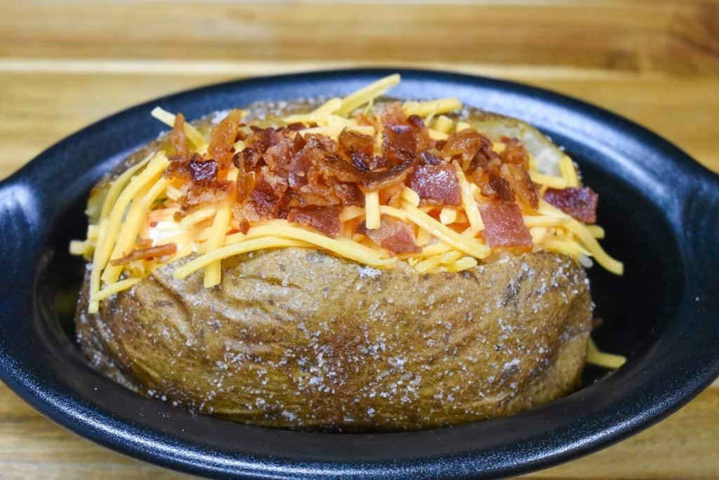 A baked potato with shredded cheese and chopped bacon on top, served in a small black crock.