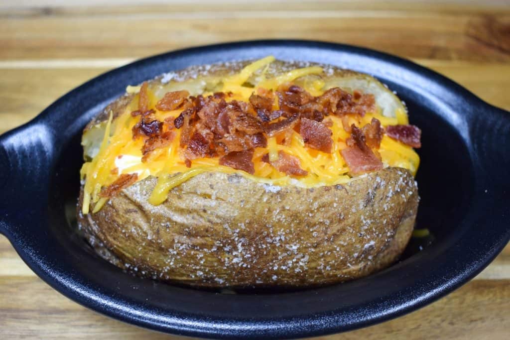 A baked potato with melted shredded cheese and chopped bacon on top, served in a small black crock.