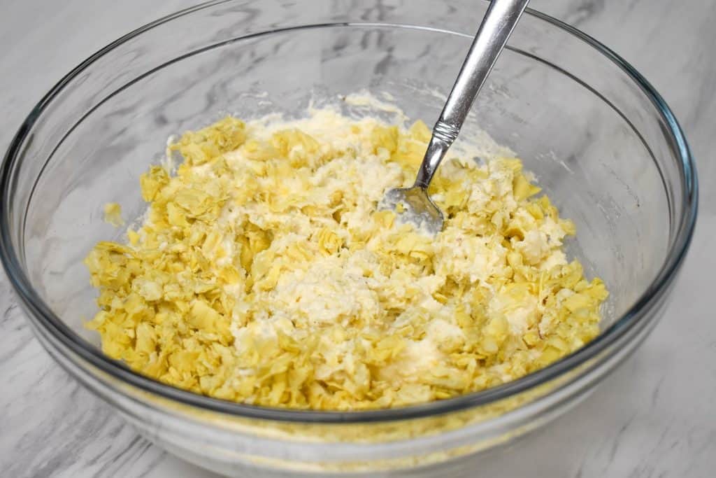 Chopped artichokes being combined with cream cheese and mayonnaise in a large, glass bowl.