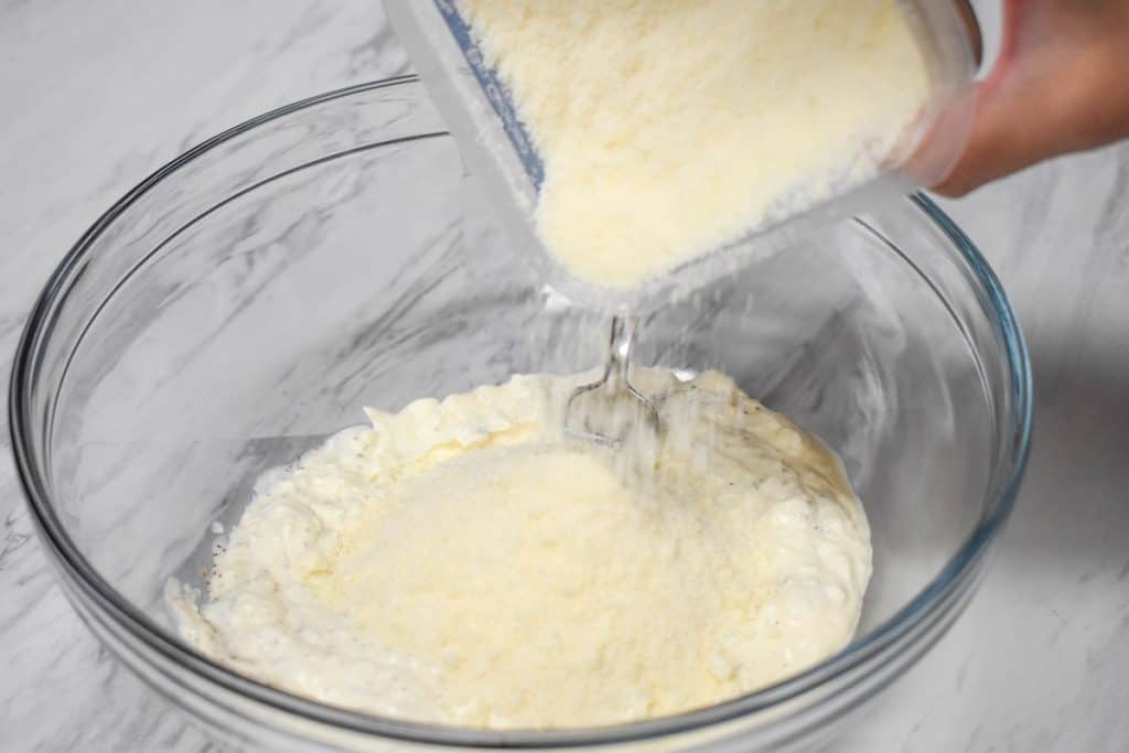 Parmesan cheese being added to cream cheese and mayonnaise in a large, glass bowl.