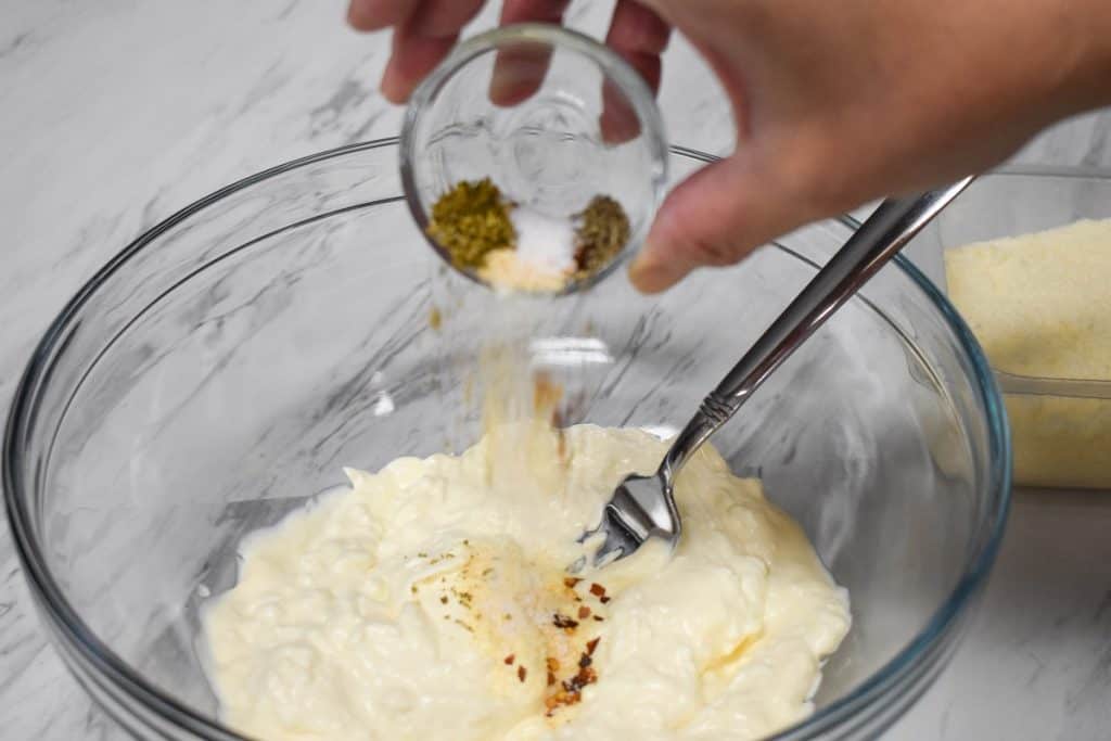 Spices being added to a large glass bowl with cream cheese and mayonnaise.