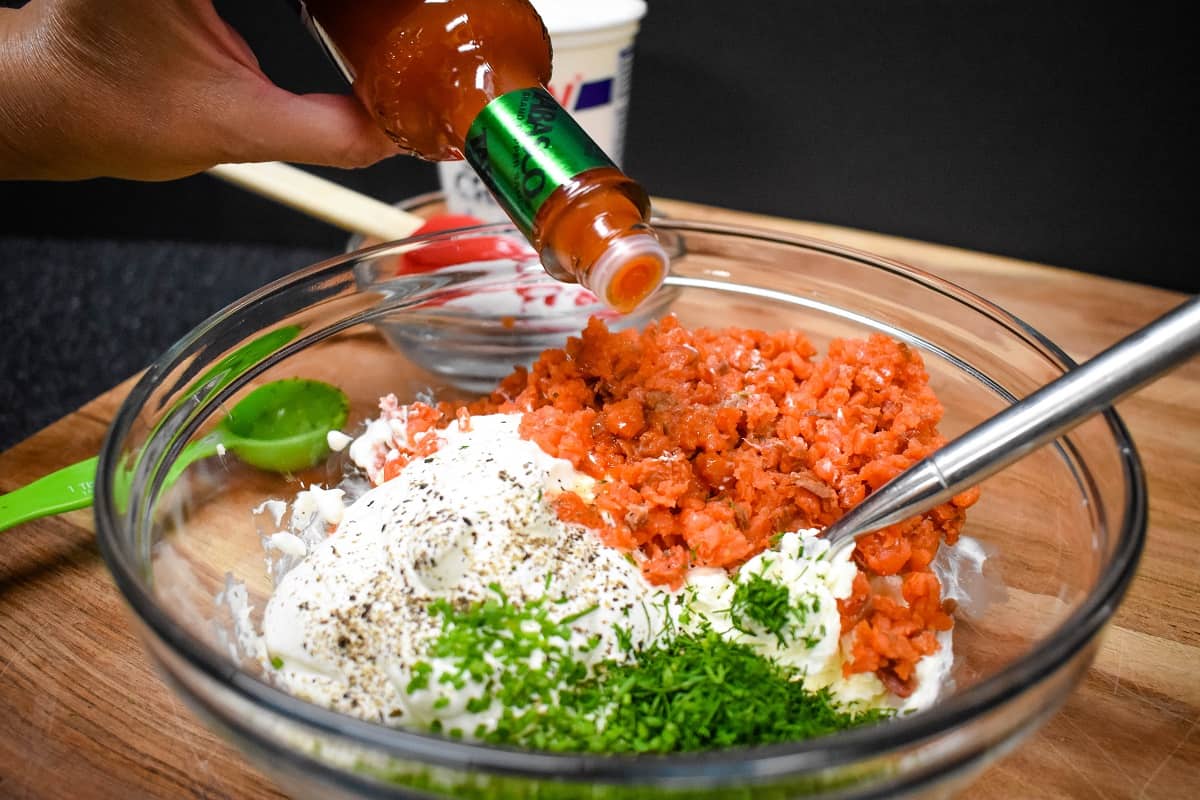 The ingredients for the salmon dip in a large, clear bowl with drops of hot sauce being added.