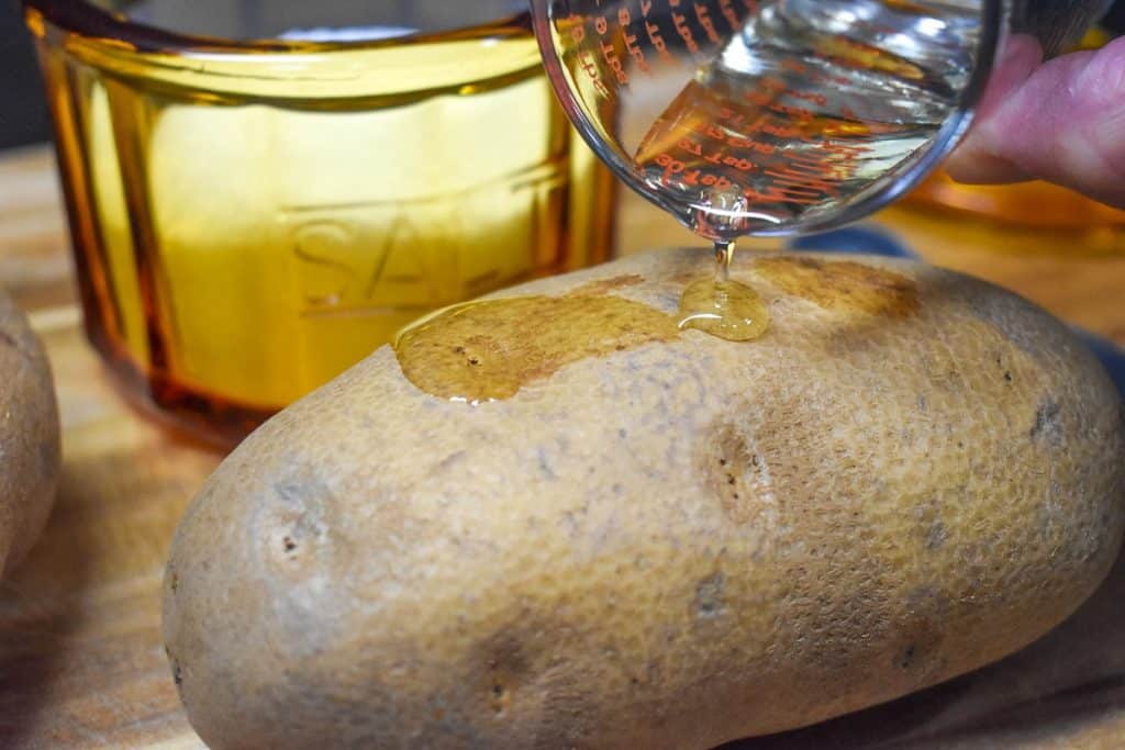 A small amount of oil being poured on a large russet potato, with an amber colored salt cellar in the background.