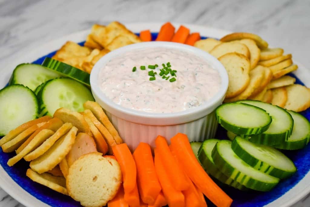Salmon Dip served in a white crock on a blue plate surrounded by bagel chips, carrot sticks and sliced cucumbers.
