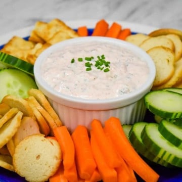 Salmon Dip served in a white crock on a blue plate surrounded by bagel chips, carrot sticks and sliced cucumbers.