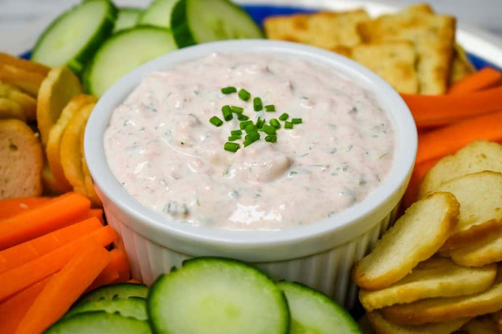 A close up picture of salmon dip in a white crock surrounded by bagel chips, carrots and cucumbers.