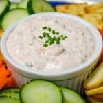 A close up picture of salmon dip in a white crock surrounded by bagel chips, carrots and cucumbers.