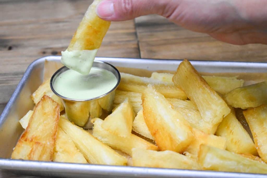 Yuca frita served in a small metal sheet pan with one piece of yuca dipped in a light green cilantro sauce.