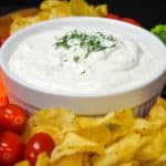 Dill dip served in a large white bowl with chips, grape tomatoes, carrots and broccoli florets arranged around the bowl.