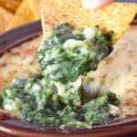 Creamy spinach and cheese being scooped out of a brown crock with a corn tortilla chip.