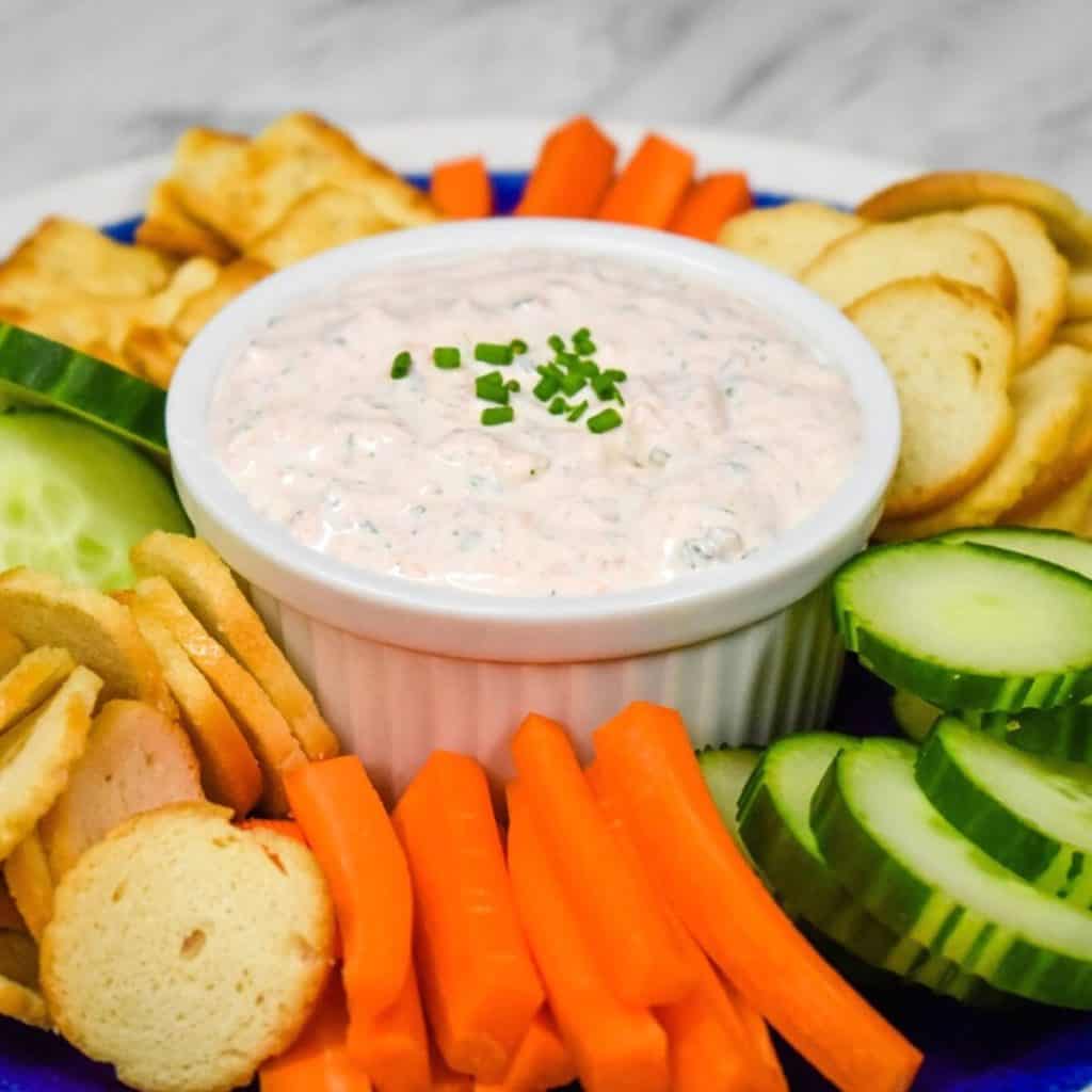 Salmon Dip served in a white crock on a blue plate surrounded by bagel chips, carrot sticks and sliced cucumbers.