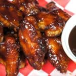 Sticky chicken wings glazed in barbecue sauce on a red and white checkered liner, with a bowl of sauce on the side.