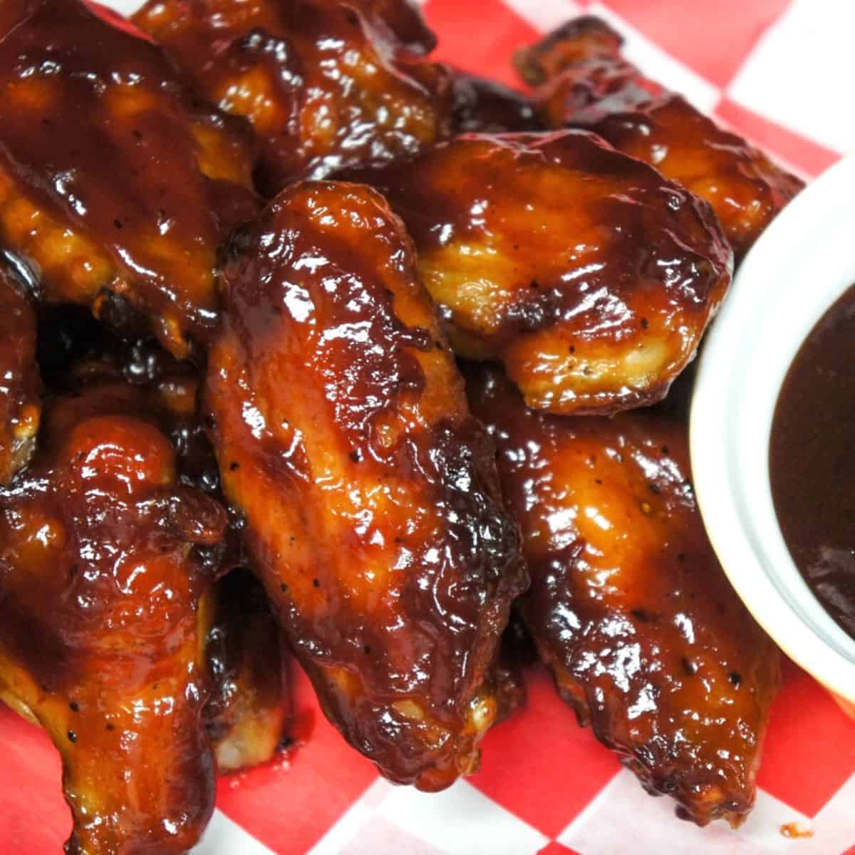 Sticky chicken wings served on a red and white checkered liner and a side of barbecue sauce.