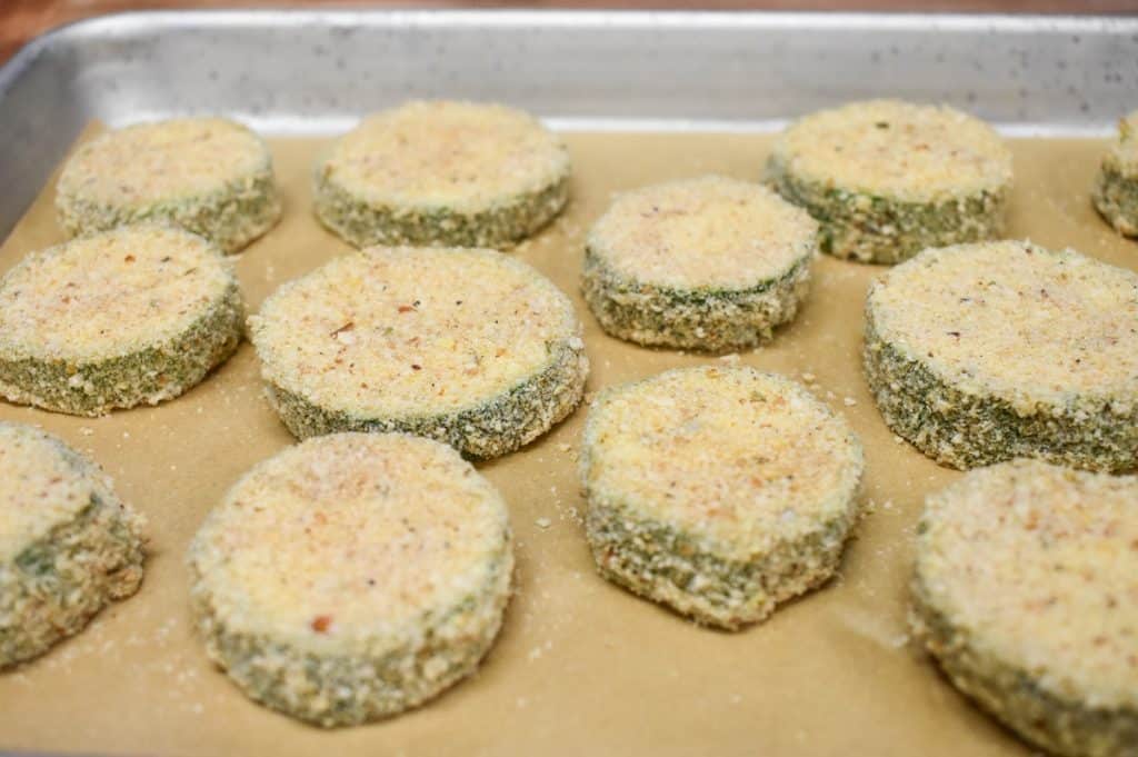 Breaded zucchini rounds arranged on a baking sheet lined with parchment paper. Before frying.