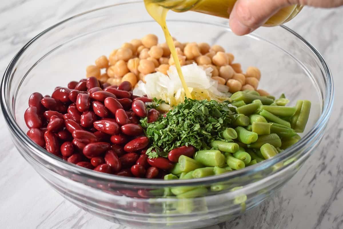 Dressing being added to red kidney beans, garbanzo beans, green beans, sliced onions and chopped parsley in a large, glass bowl.